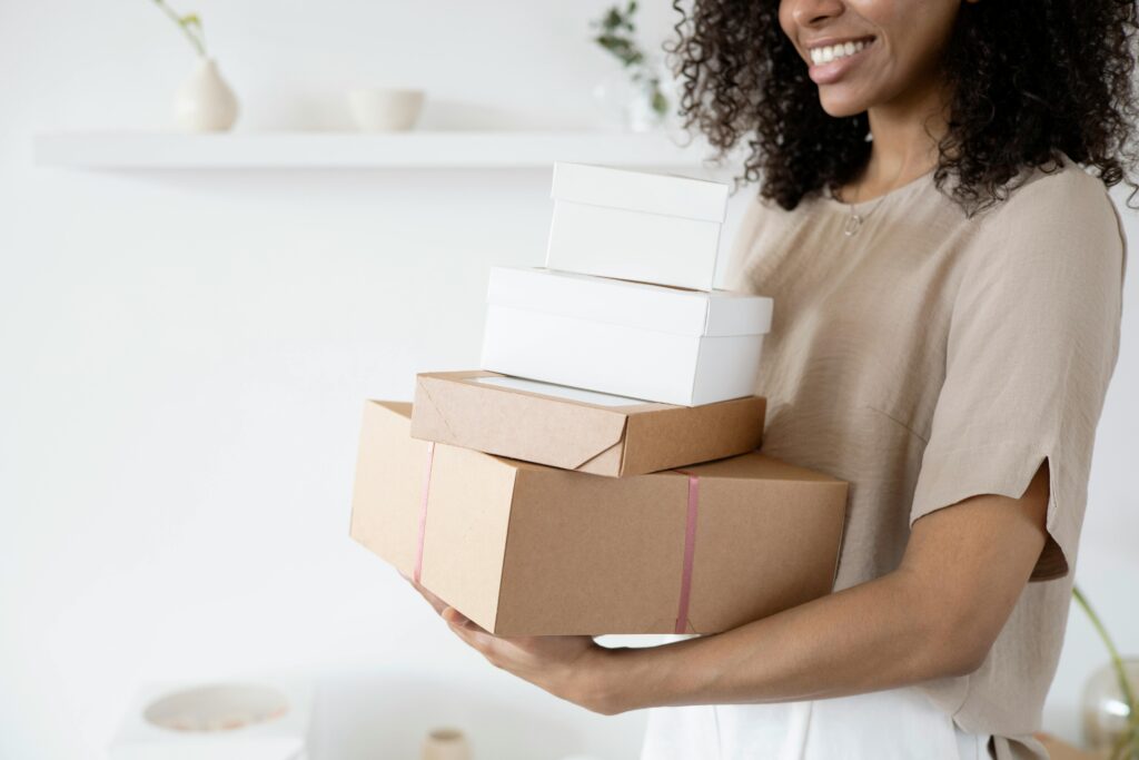 Woman with curly hair carrying stack of packaging boxes in a bright room.
